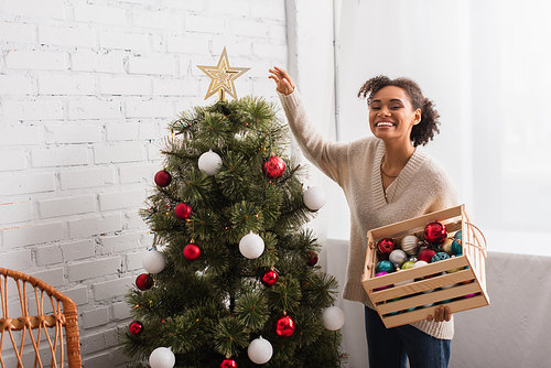 Cheerful african american woman holding box with christmas balls near tree at home