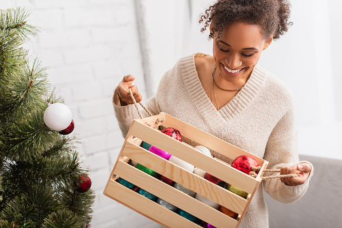 Smiling african american woman holding wooden box with balls near christmas tree at home