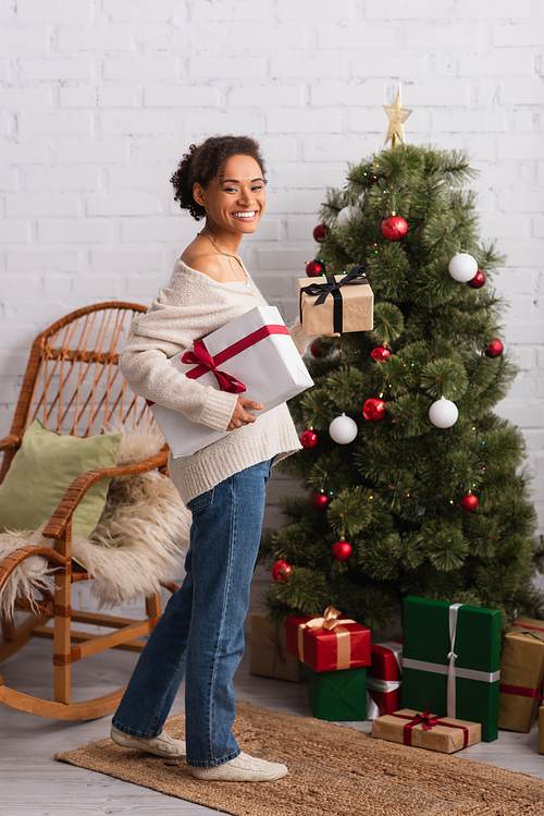 Smiling african american woman looking at camera while holding presents near christmas tree at home