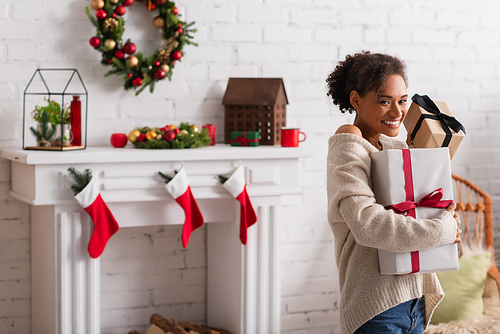Cheerful african american woman holding christmas presents near blurred fireplace at home
