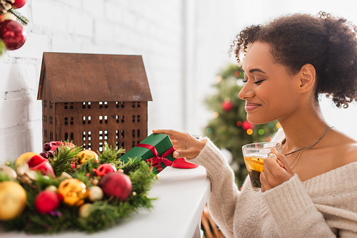 Smiling african american woman holding tea and gift on fireplace with christmas wreath