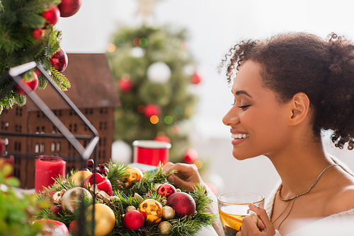 Side view of smiling african american woman holding cup of tea near decorated christmas wreath