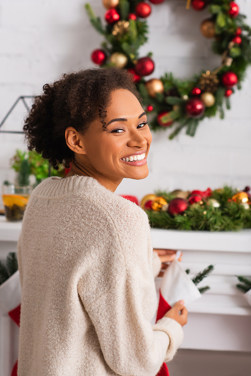 Young african american woman smiling at camera near blurred fireplace with christmas wreath