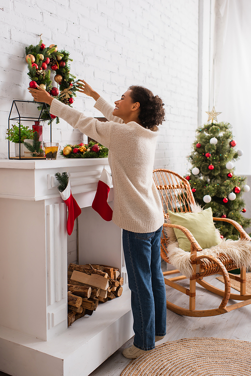 Side view of positive african american woman holding christmas wreath near decorated fireplace at home