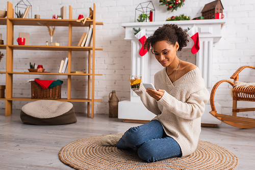 Young african american woman with cup of tea using smartphone near decorated fireplace at home