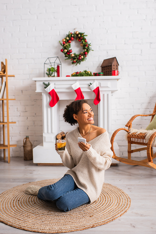 Happy african american woman holding tea and smartphone near decorated fireplace during christmas