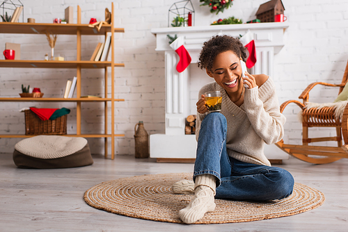 Smiling african american woman with cup of tea talking on cellphone near decorated fireplace at home