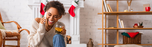 Cheerful african american woman talking on smartphone and holding tea near decorated fireplace, banner
