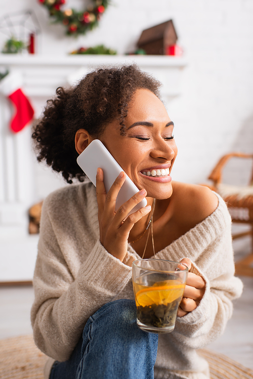Cheerful african american woman talking on smartphone and holding cup of tea at home