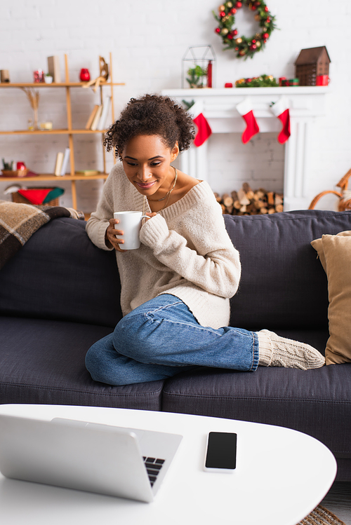 Smiling african american woman with cup looking at laptop during christmas at home