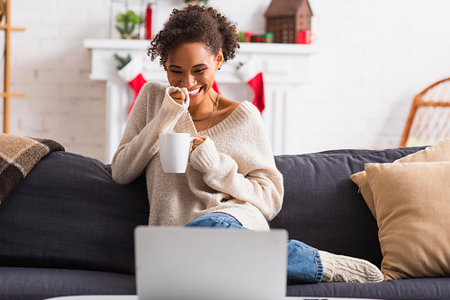 Cheerful african american woman with cup looking at blurred laptop during christmas at home