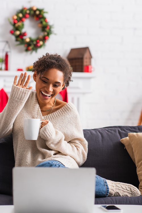 Smiling african american woman with cup having video call on laptop during christmas