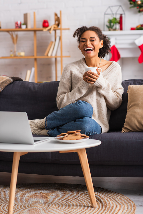 Laughing african american woman holding cup near laptop and christmas cookies at home