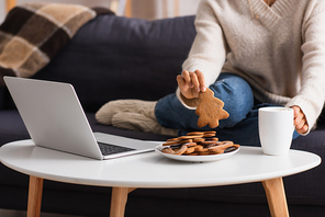 Cropped view of african american woman taking christmas cookie near cup and laptop at home