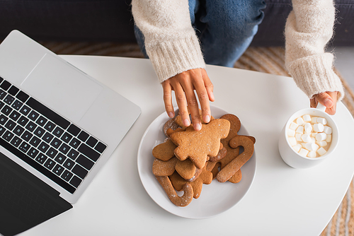Top view of african american woman taking christmas cookie near cup of cocoa with marshmallows and laptop at home