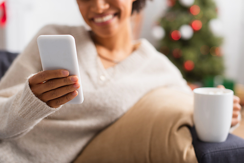 Cropped view of smartphone in hand of blurred african american woman with cup at home