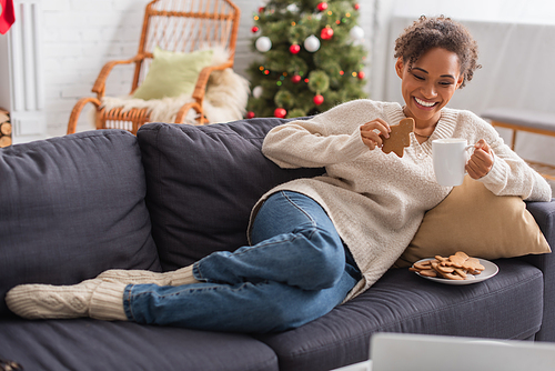 Positive african american woman with christmas gingerbread and cup looking at blurred laptop at home