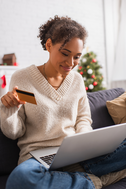 Smiling african american woman holding credit card and using laptop during christmas at home