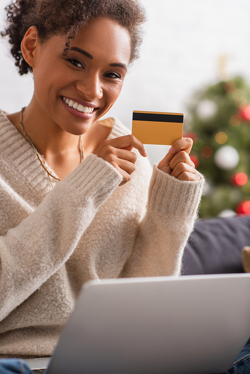 Smiling african american woman holding credit card near blurred laptop during christmas at home