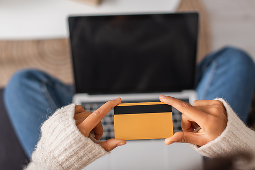 Cropped view of african american woman holding credit card near blurred laptop at home
