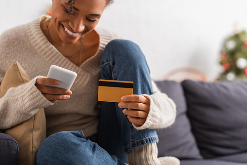 Positive african american woman using smartphone and credit card at home during christmas