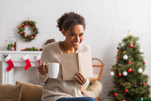 Smiling african american woman with cup and book looking at camera during christmas at home