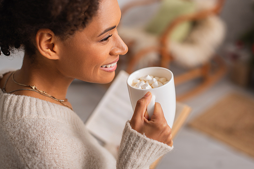 Side view of happy african american woman holding cup of cocoa with marshmallows and blurred book at home