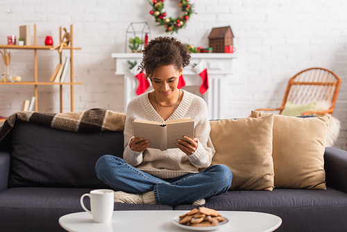 Smiling african american woman reading book near christmas cookies and cup at home