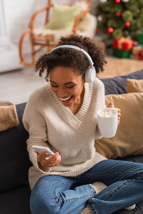 Cheerful african american woman in headphones holding cocoa and smartphone at home