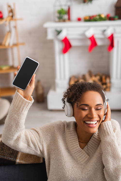 Smiling african american woman in headphones holding smartphone with blank screen during christmas at home