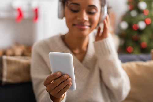 Blurred african american woman in headphones using smartphone during christmas at home