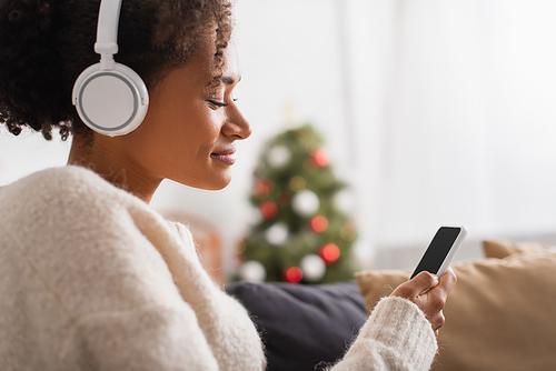 Side view of african american woman in headphones using cellphone during christmas at home