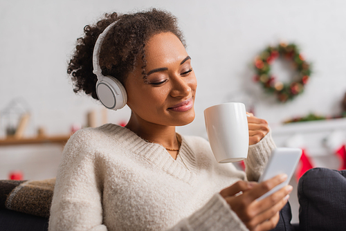 Smiling african american woman in headphones holding cup and cellphone during christmas at home