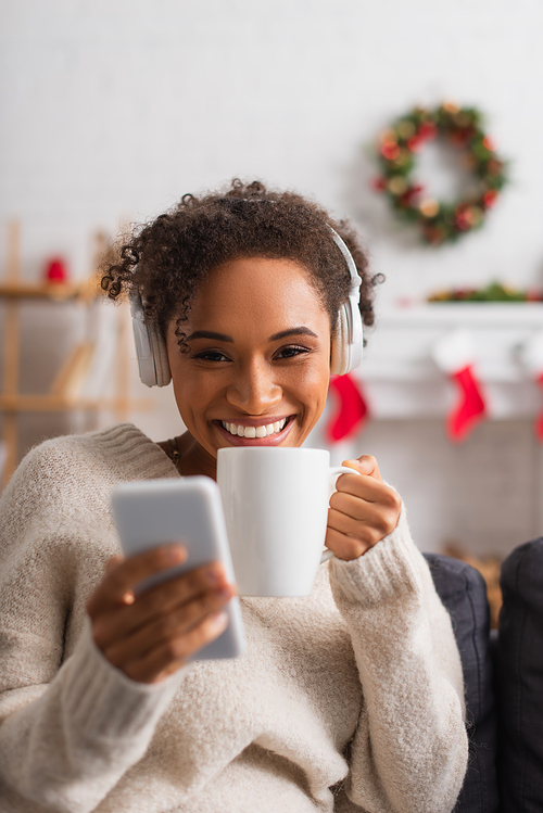 Smiling african american woman in headphones holding cup and blurred smartphone during christmas at home