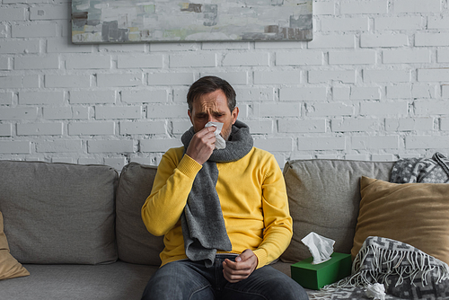 sick man in warm scarf sitting on sofa at home and sneezing in paper napkin