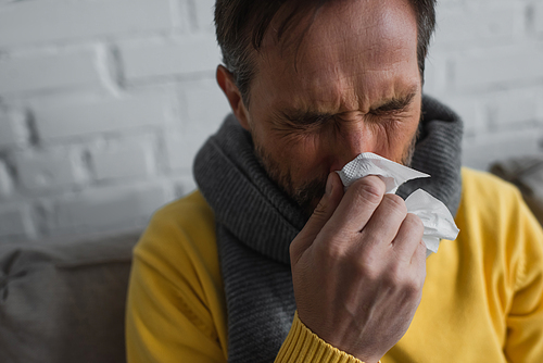 sick man in warm scarf holding paper napkin and sneezing with closed eyes