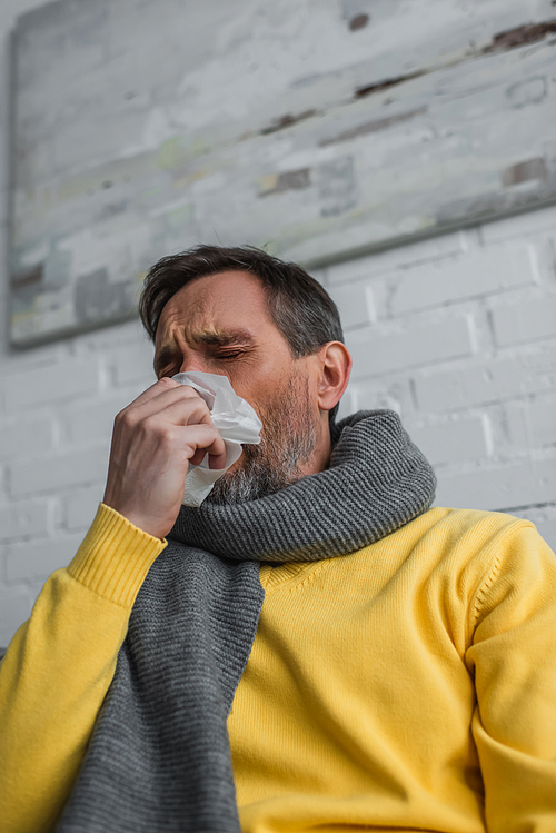 low angle view of diseased man in warm scarf holding paper napkin and sneezing with closed eyes