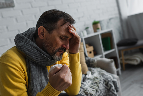 diseased man in warm scarf holding paper napkin and suffering from headache