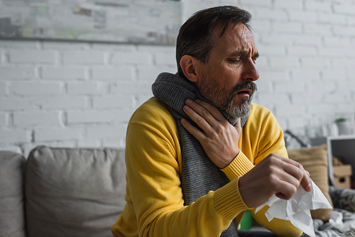 sick man in warm scarf touching sore throat and coughing while holding paper napkin