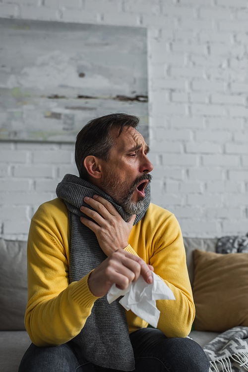 sick man in warm scarf holding paper napkin and coughing on couch