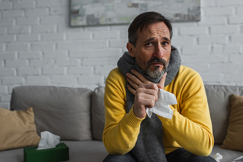 diseased man in warm scarf touching sore throat while holding paper napkin and looking at camera