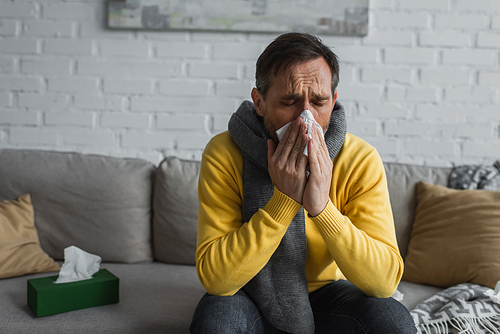 ill man in warm scarf sneezing in paper napkin with closed eyes while sitting on couch