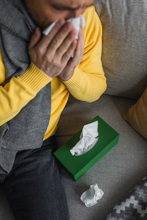 diseased man sneezing while sitting on couch near pack of paper napkins