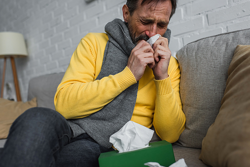 sick man in warm scarf sneezing in paper napkin with closed eyes on couch