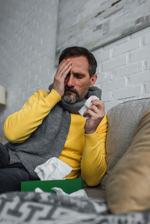 diseased man obscuring face with hand while sitting on couch with paper napkin and suffering from headache