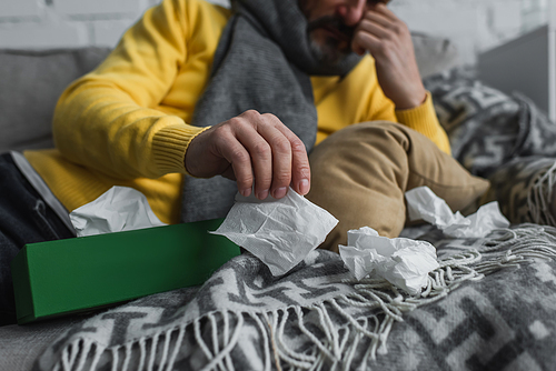 partial view of sick man lying on couch with warm blanket and holding paper napkin