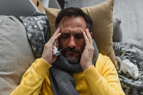 top view of sick man in warm scarf touching head while lying on couch and suffering from migraine