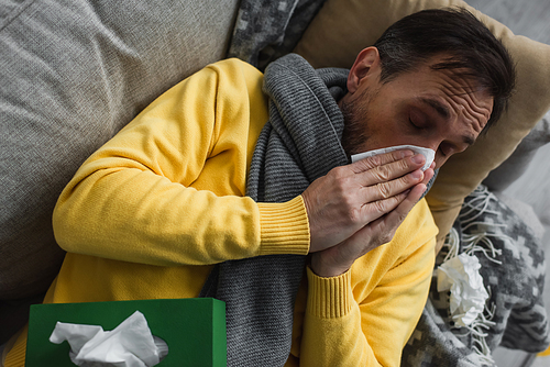 top view of sick man in warm scarf lying on sofa and sneezing in paper napkin