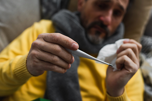 selective focus of electronic thermometer in hand on sick man suffering from fever on blurred background