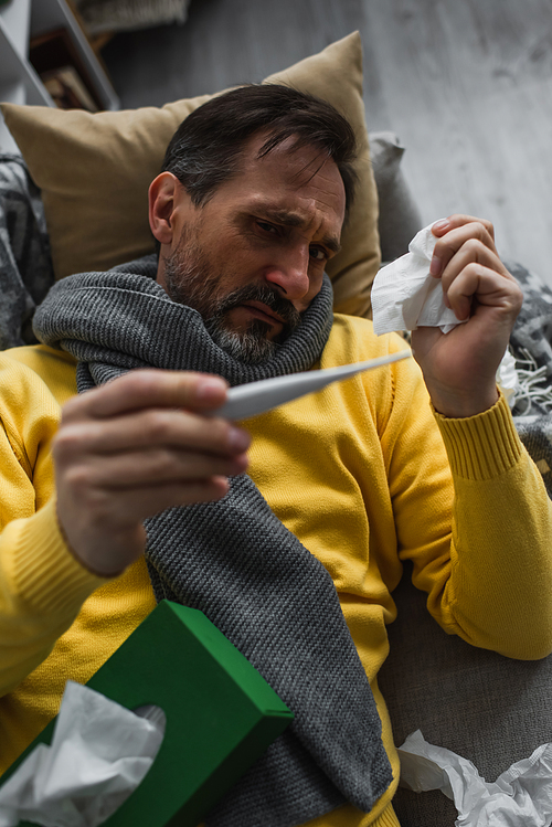 sick man in warm scarf holding paper napkin and looking at electronic thermometer